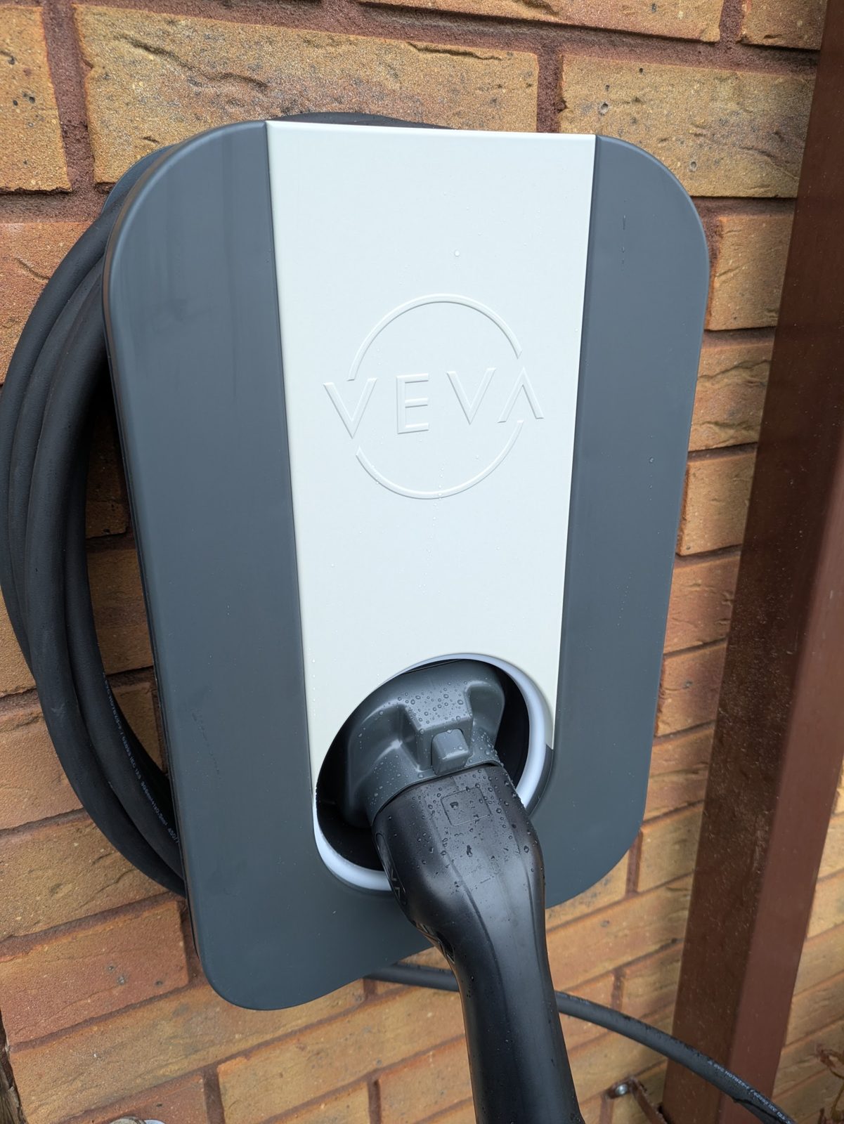 A white Veva EV charger with its cable plugged in, on a brick wall